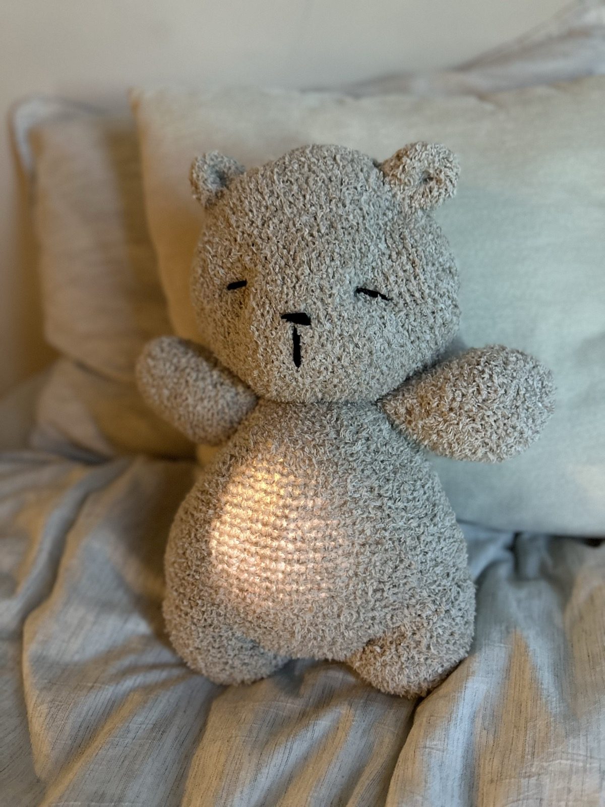 A grey knit teddy bear sitting on a bed, its belly glowing warm from inside