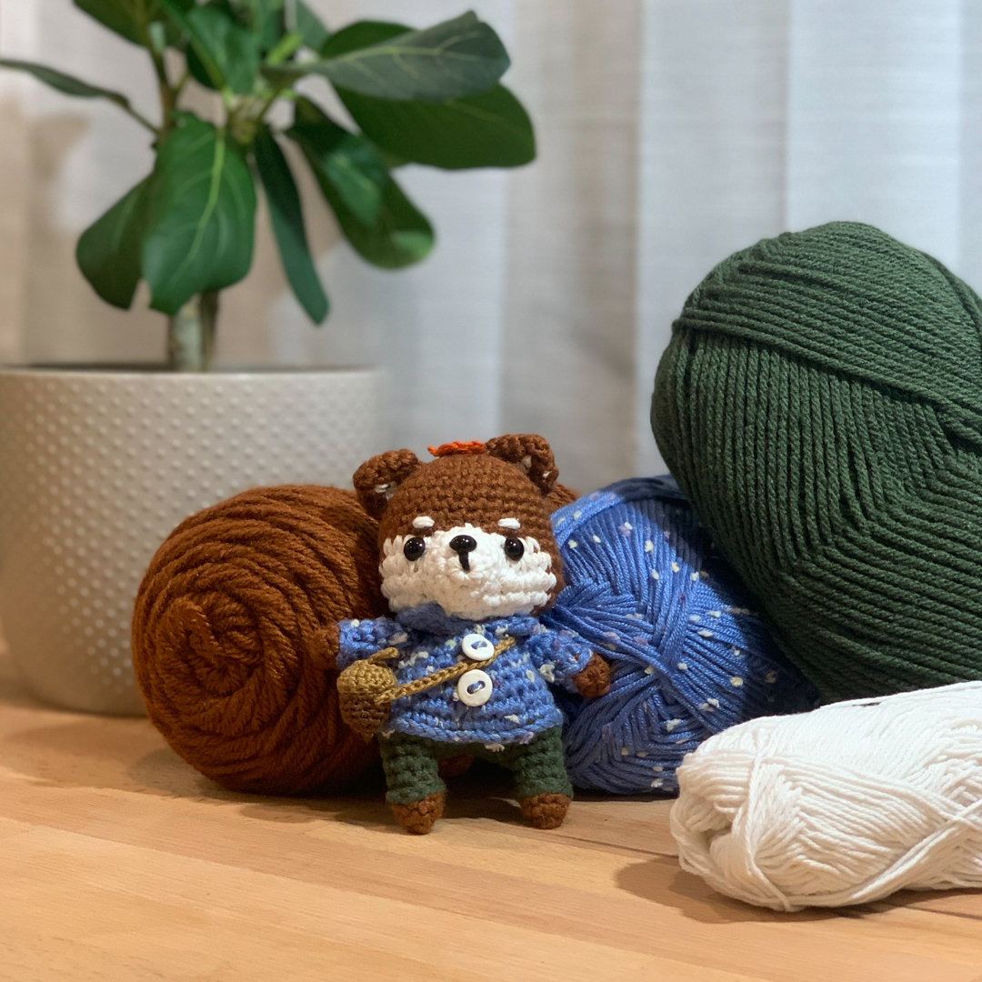 Toby the Shiba, the finished amigurumi in a blue coat, beside balls of yarn